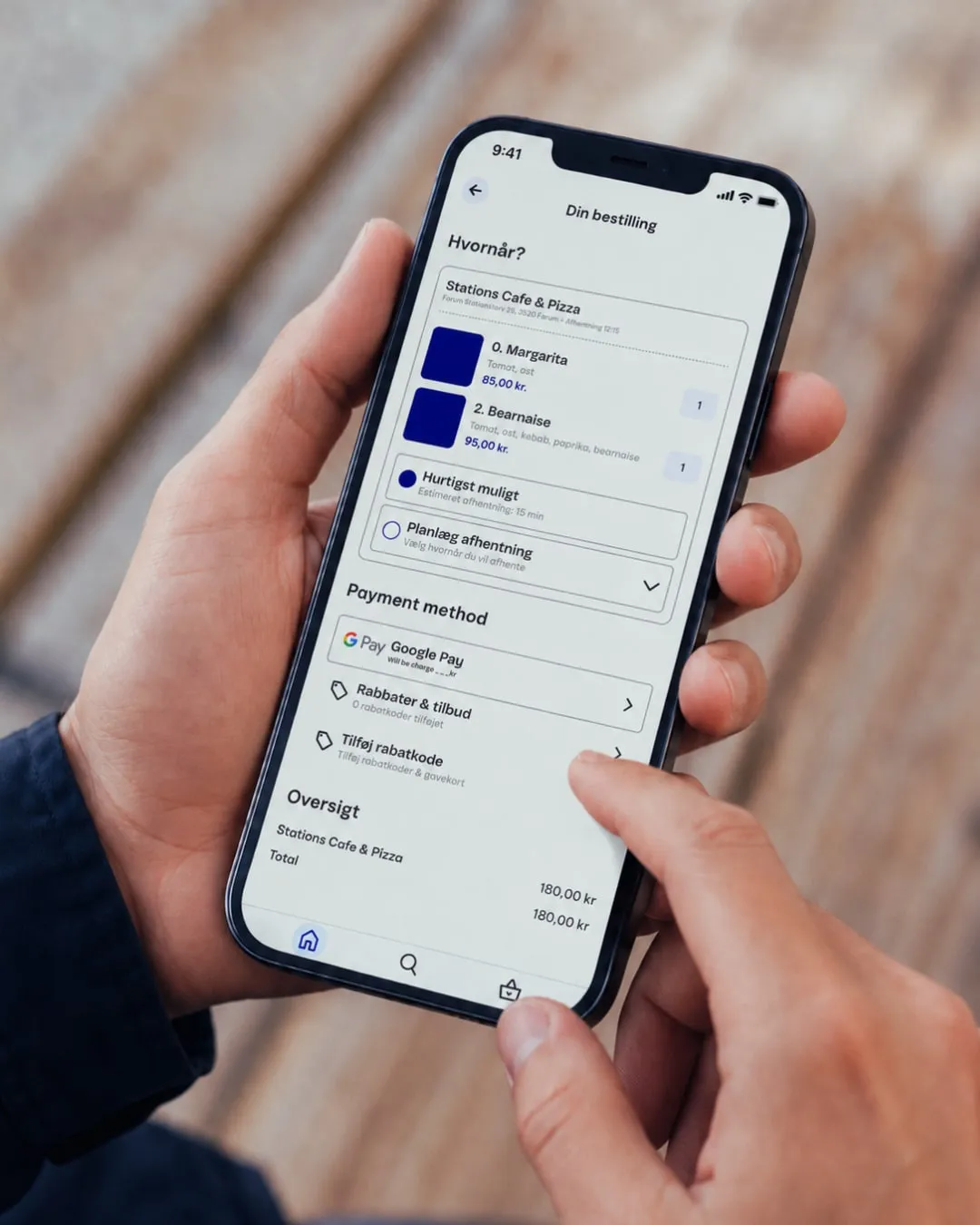 Hænder holder en telefon med E-BIZ-appens bestillingsskærm: Din bestilling, valg af afhentningstid og betaling.