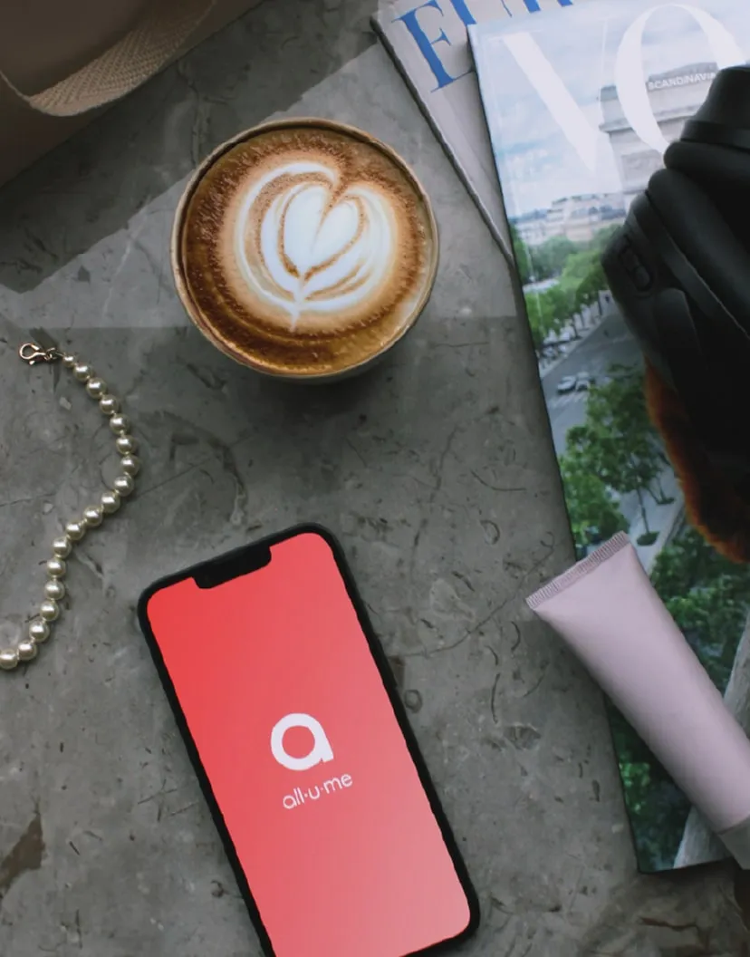 Telefon med all.u.me-logoet på skærmen ved siden af en kaffe, en perlekæde og magasiner.