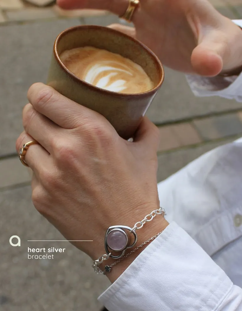 Hænder om en kop kaffe. Den ene bærer et sølvarmbånd med lyserød sten. Tekst: heart silver bracelet.