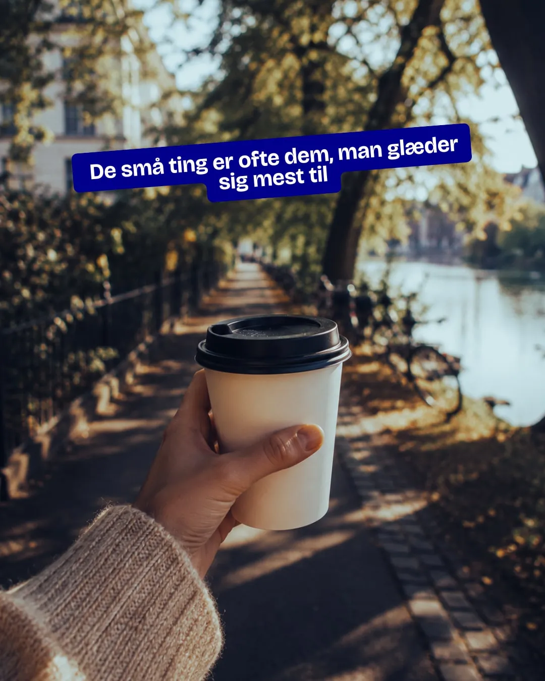 Hånd holder en kaffe to go på en solrig sti ved søerne. Tekst: De små ting er ofte dem, man glæder sig mest til.